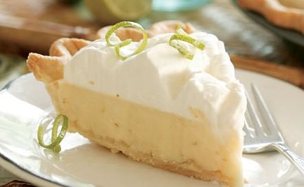 A slice of creamy Key Lime Pie made with authentic Key West key limes and a buttery digestive biscuit crust is topped with whipped cream and garnished with lime zest curls on a white plate. A fork rests beside the pie slice. The background is slightly out of focus, featuring a lime and other blurred elements.